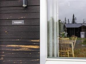 A wooden exterior wall with a sign reading 'TJÄDERN' and a window reflecting a wooden table and chairs.