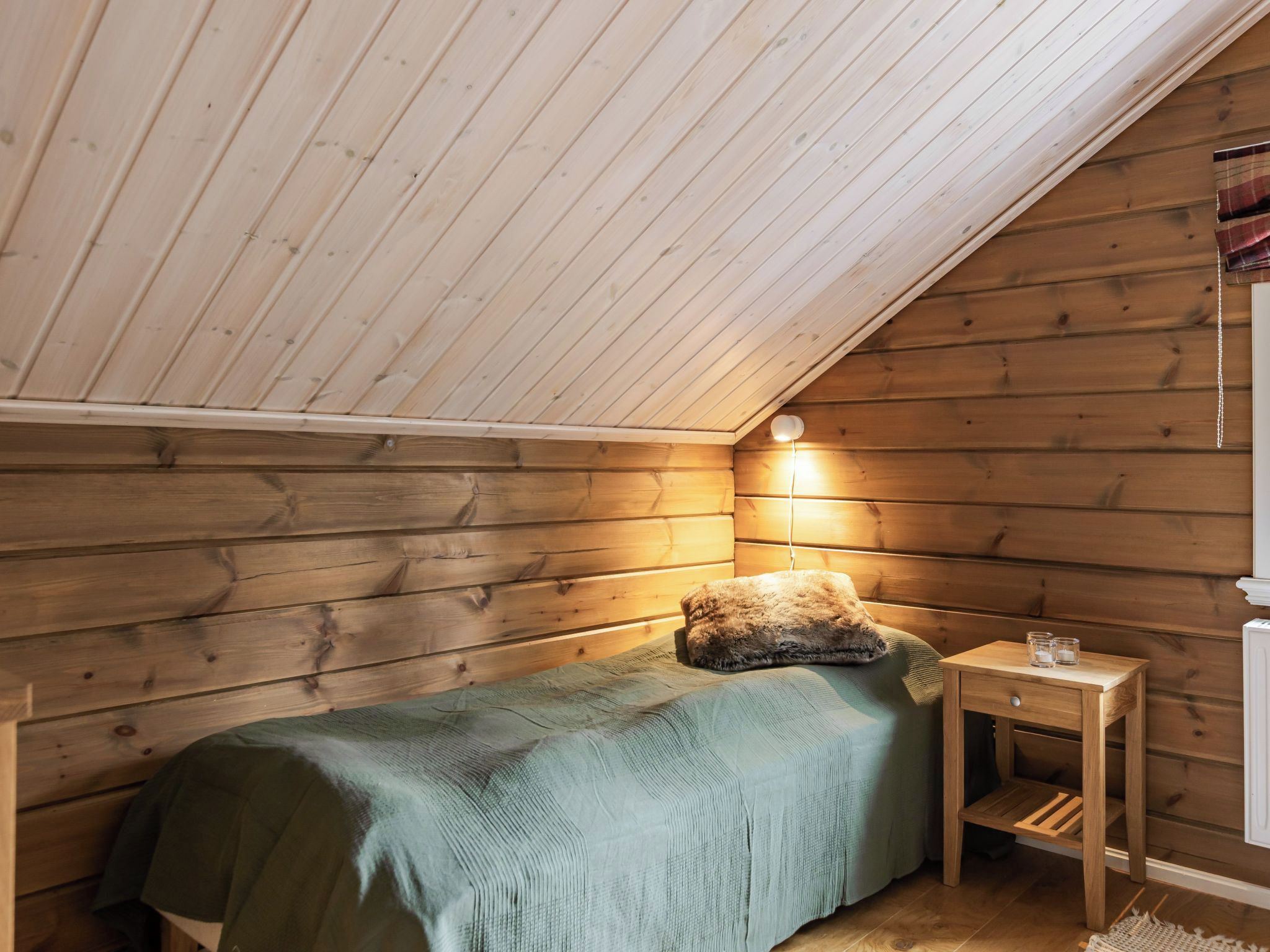Une petite pièce chaleureuse avec un plafond en pente, des murs en bois, un lit simple recouvert d’une couverture verte, une petite table de chevet avec une lampe et un radiateur.