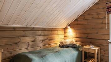 Une petite pièce chaleureuse avec un plafond en pente, des murs en bois, un lit simple recouvert d’une couverture verte, une petite table de chevet avec une lampe et un radiateur.