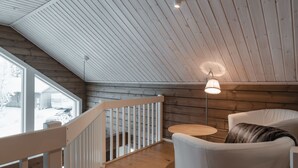 Interior de una cabaña de madera con techo inclinado, barandilla blanca, mesa redonda y sofá con un cojín decorativo.
