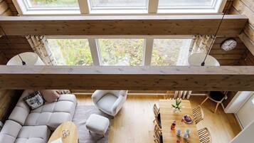 Une pièce aux hauts plafonds, avec un plafond en bois, un canapé, une table à manger avec des chaises, et une vue dégagée grâce à de grandes fenêtres.