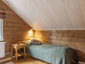 A cozy attic bedroom with a sloped ceiling, wooden walls, a single bed with a green blanket, a bedside table with a fruit bowl, and a wall-mounted light.