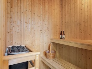 Een sauna met houten wanden, een bankje, een schaal met stenen, een houten emmer en twee flessen op een plank.