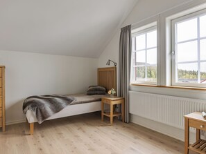 Une petite chambre avec un lit simple, une tête de lit en bois, une table de chevet avec une lampe, une fenêtre avec des rideaux et un radiateur.