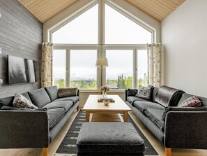 Een moderne woonkamer met twee grijze banken, een houten salontafel en een groot raam met uitzicht op bomen en bergen.