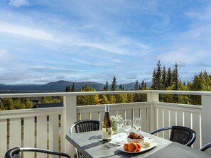 Une table dressée pour deux personnes, avec une bouteille de vin, des verres, une assiette de nourriture et une vue panoramique sur les montagnes et les arbres.