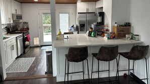 Eine moderne Küche mit weißen Schränken, einem Edelstahlkühlschrank, einer Mikrowelle und einer Kaffeemaschine. An der Kücheninsel stehen drei Barhocker und ein Mülleimer befindet sich in der Nähe der Tür.