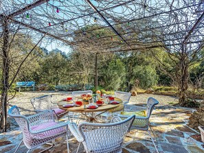 Un área de comedor al aire libre con sillas de mimbre y una mesa redonda dispuesta para una comida, rodeada de árboles y un suelo empedrado de piedra.