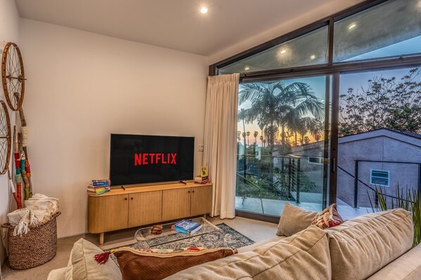 Un salon moderne avec une grande télévision affichant le logo « NETFLIX », une console en bois sur laquelle sont posés des livres, un panier en osier et une table basse en verre.