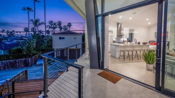 A modern kitchen with a glass door leading to a balcony overlooking palm trees and a sunset.