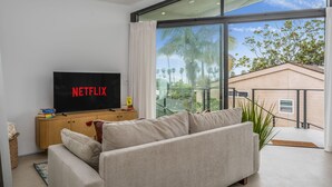 A modern living room with a large flat-screen TV, a sofa, and a view of palm trees through large windows.