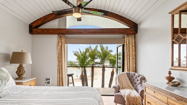 A bedroom with a large bed, a wooden dresser, a chair, and a balcony with a view of palm trees. - New! Bayfront 3 BD/3BA Retreat with Private Patio! (San Diego)