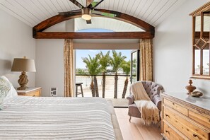 Un dormitorio con una cama grande, un aparador de madera, una silla y un balcón con vistas a palmeras.