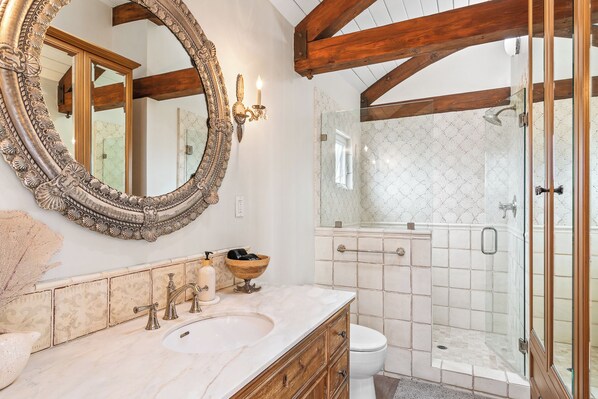 A bathroom with a large ornate mirror, a marble sink, wooden cabinetry, a white toilet, and a glass-enclosed shower area.