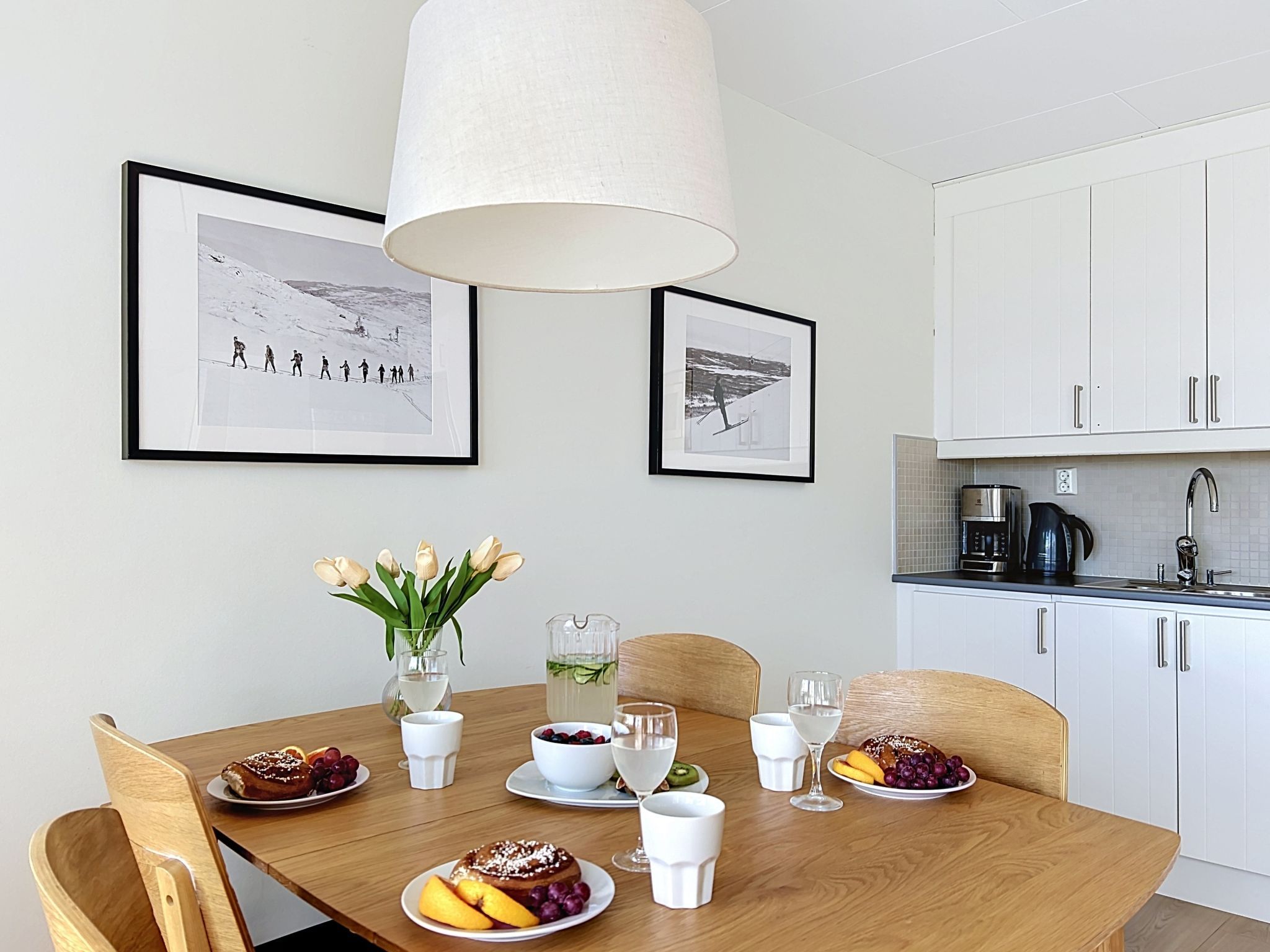 En matplats med ett träbord dukat för en måltid, två inramade fotografier på väggen och ett kök i bakgrunden.