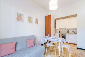 Ein modernes Wohnzimmer mit grauem Sofa, einem Esstisch mit Stühlen und einer Küchenzeile mit weißen Schränken und einer gefliesten Spritzschutzleiste.