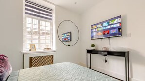 Quarto com cama, televisão na parede, janela com persianas e uma pequena mesa com uma planta.
