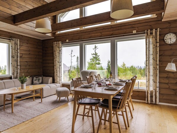 Una habitación amplia con techo de madera, ventanas grandes, una mesa de comedor lista para seis personas y un rincón de estar acogedor con sofá y sillones.