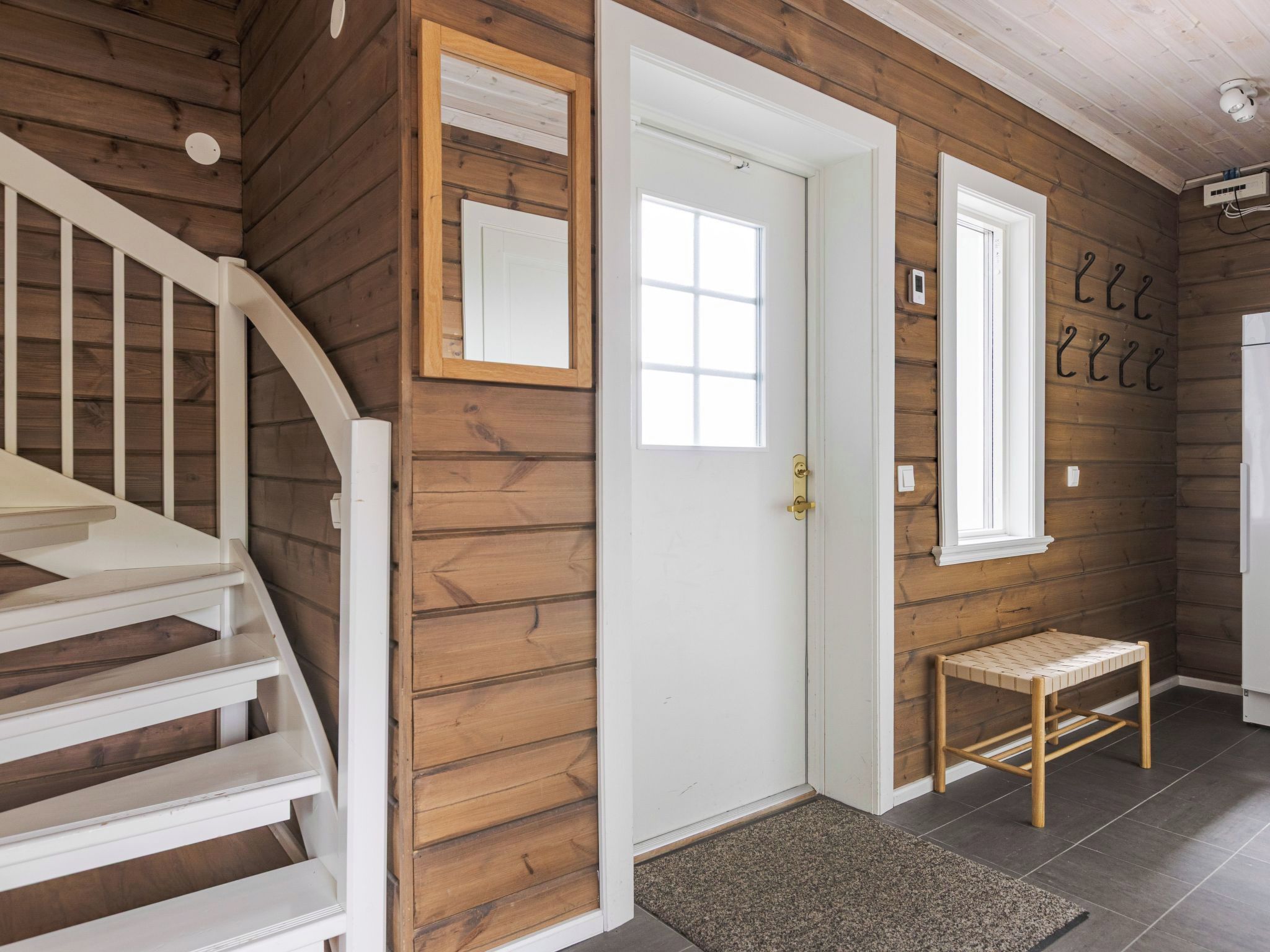 Un intérieur en bois avec un escalier blanc, une porte, un petit banc et un miroir fixé au mur.