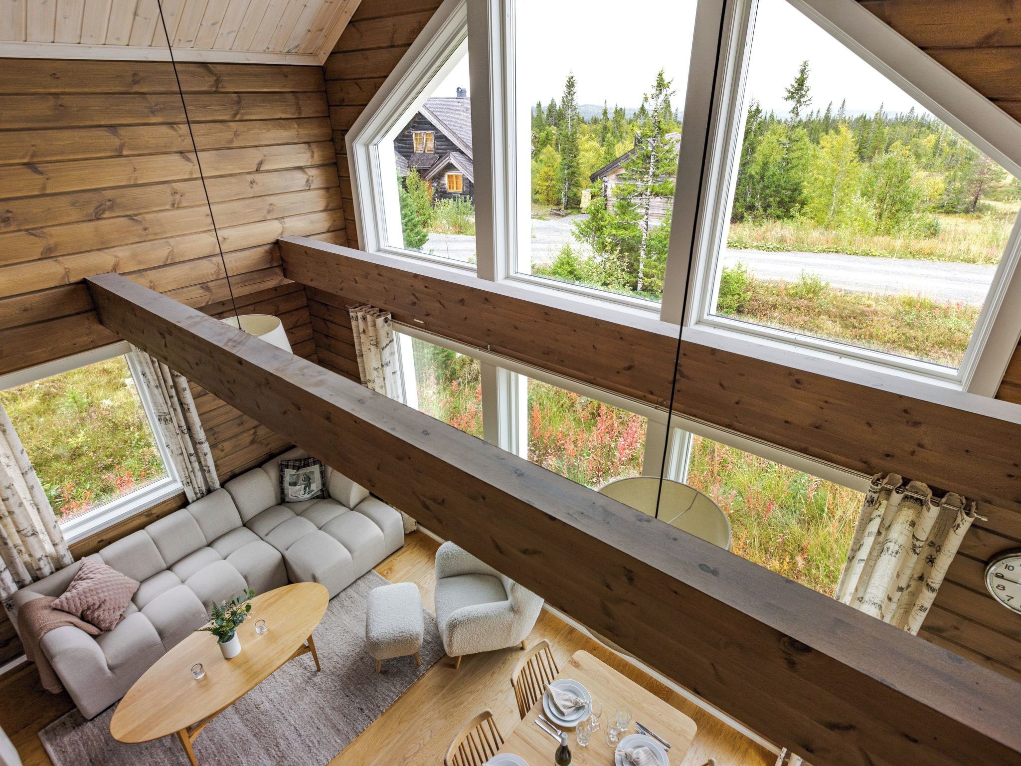 Une pièce aux hauts plafonds, avec un décor intérieur en bois, un ensemble de canapés, une table à manger et de grandes fenêtres donnant sur une zone boisée.