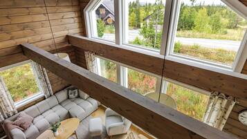 Une pièce aux hauts plafonds, avec un décor intérieur en bois, un ensemble de canapés, une table à manger et de grandes fenêtres donnant sur une zone boisée.