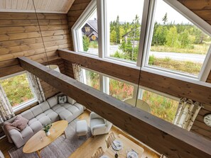A high-ceilinged room with a wooden interior, a sofa set, a dining table, and large windows offering a view of a forested area.