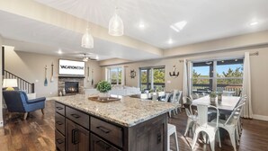 Amplia sala de estar con una isla grande, mesa de comedor y sillas. Incluye una chimenea, televisor y vistas al exterior.