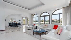 Ein großzügiger Wohnbereich mit einem weißen Sofa, einem Couchtisch und einem Esstisch mit Stühlen. Große Fenster bieten einen Blick auf die Umgebung.