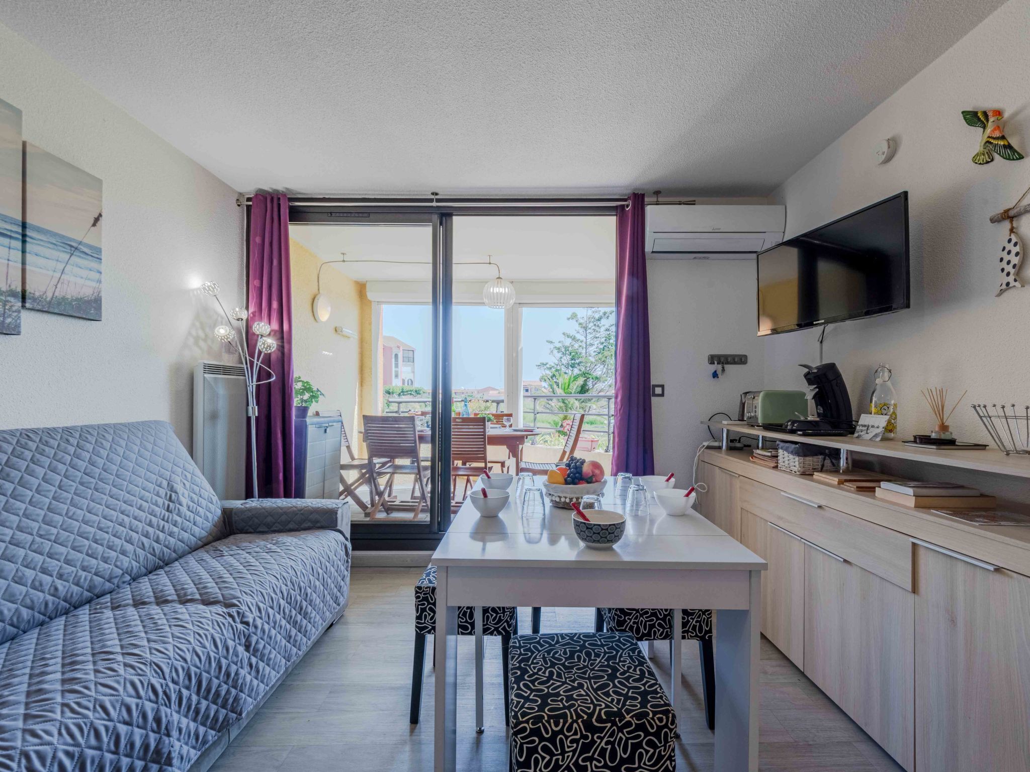 Ein modernes Wohnzimmer mit Sofa und Esstisch, eine Küchenzeile mit Fernseher und ein Balkon mit Blick.