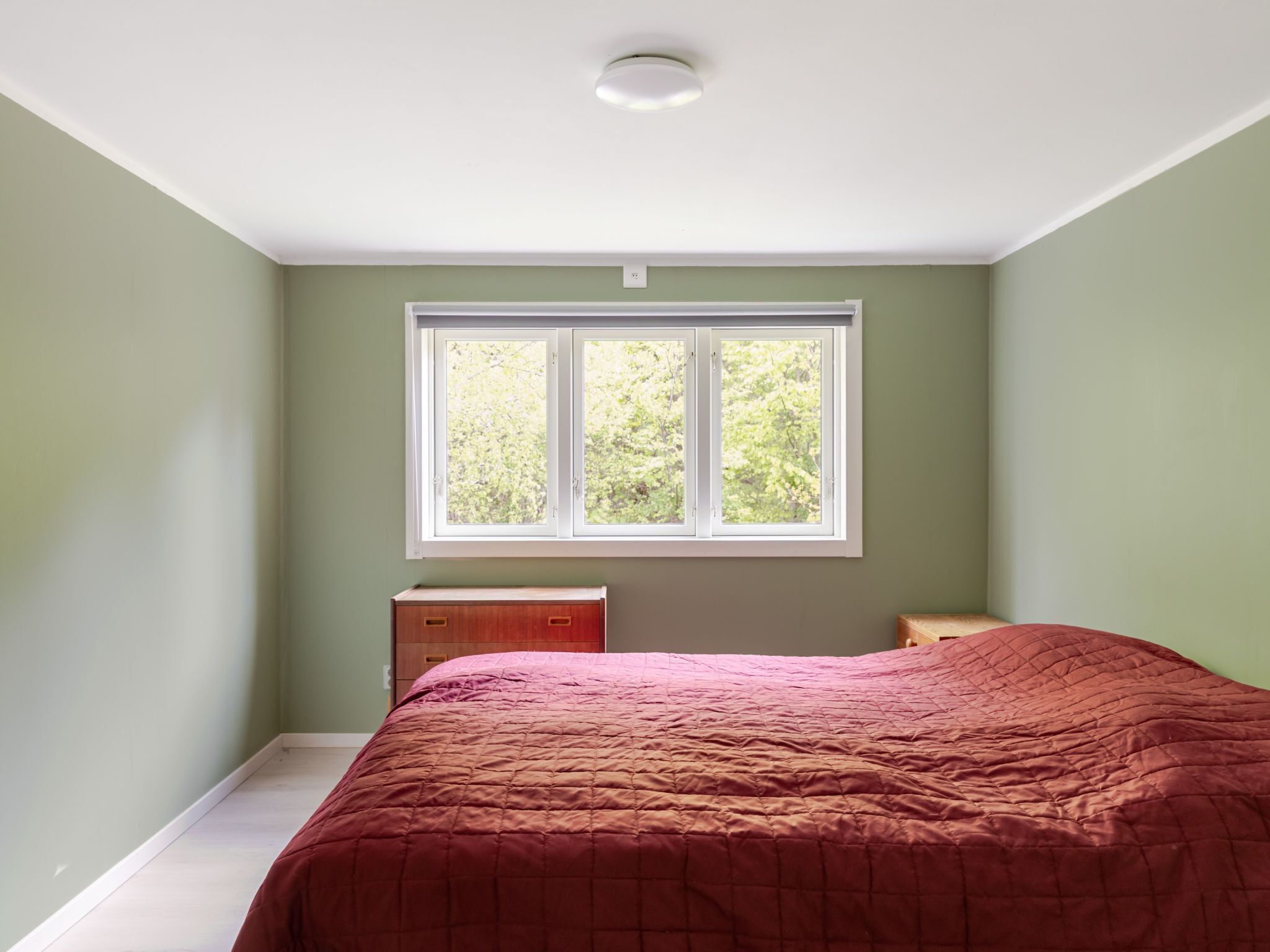 A bedroom with a large window, a bed with a red quilt, and a wooden dresser.