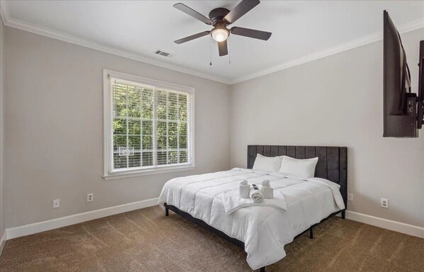 A bedroom with a bed, a ceiling fan, a window, and a wall-mounted TV.