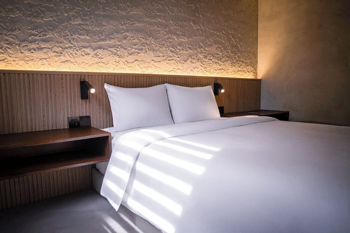 A modern bedroom with a wooden headboard, a bed with white linens, and two wall-mounted reading lamps.