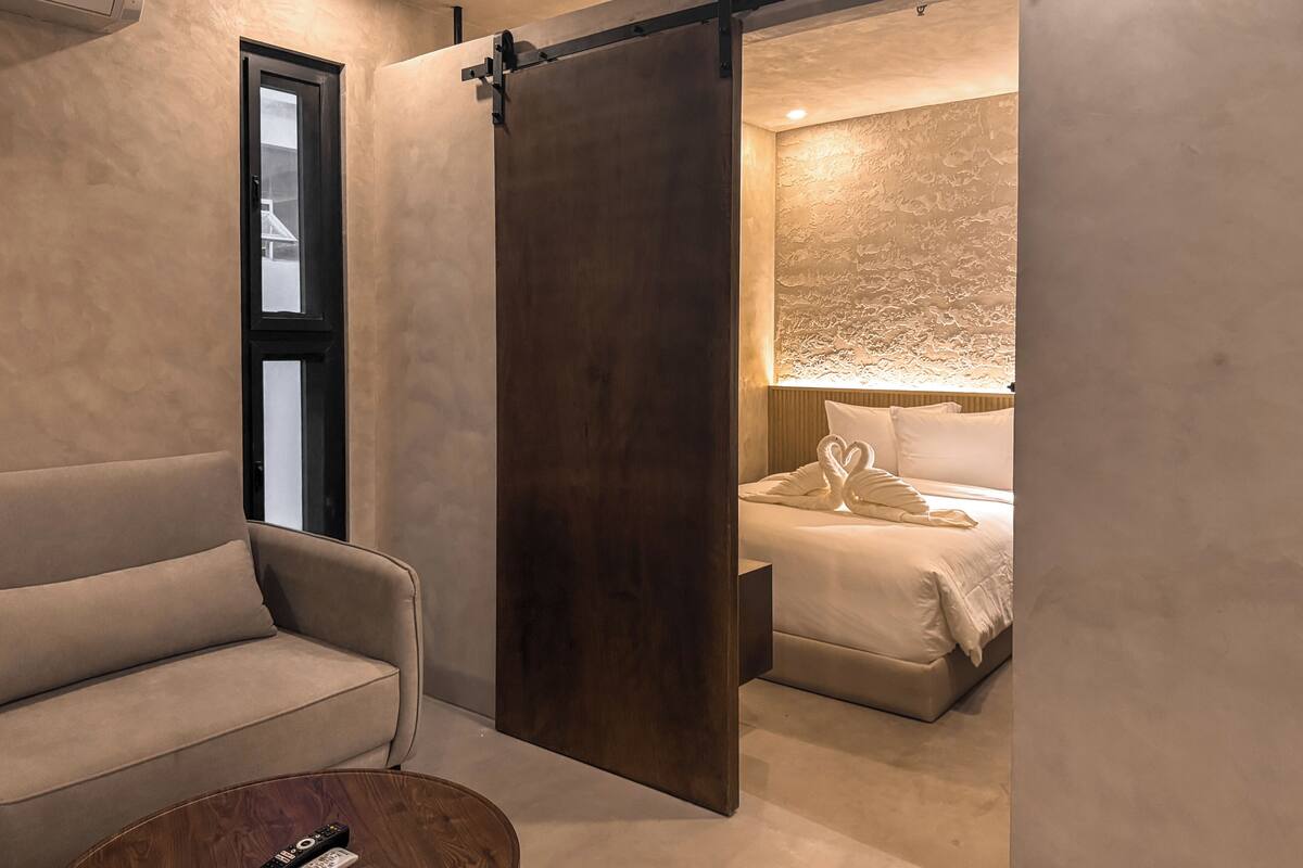 A modern hotel room with a sliding door separating the living area and bedroom. The room features a sofa, a coffee table, and a bed with neatly arranged towels.