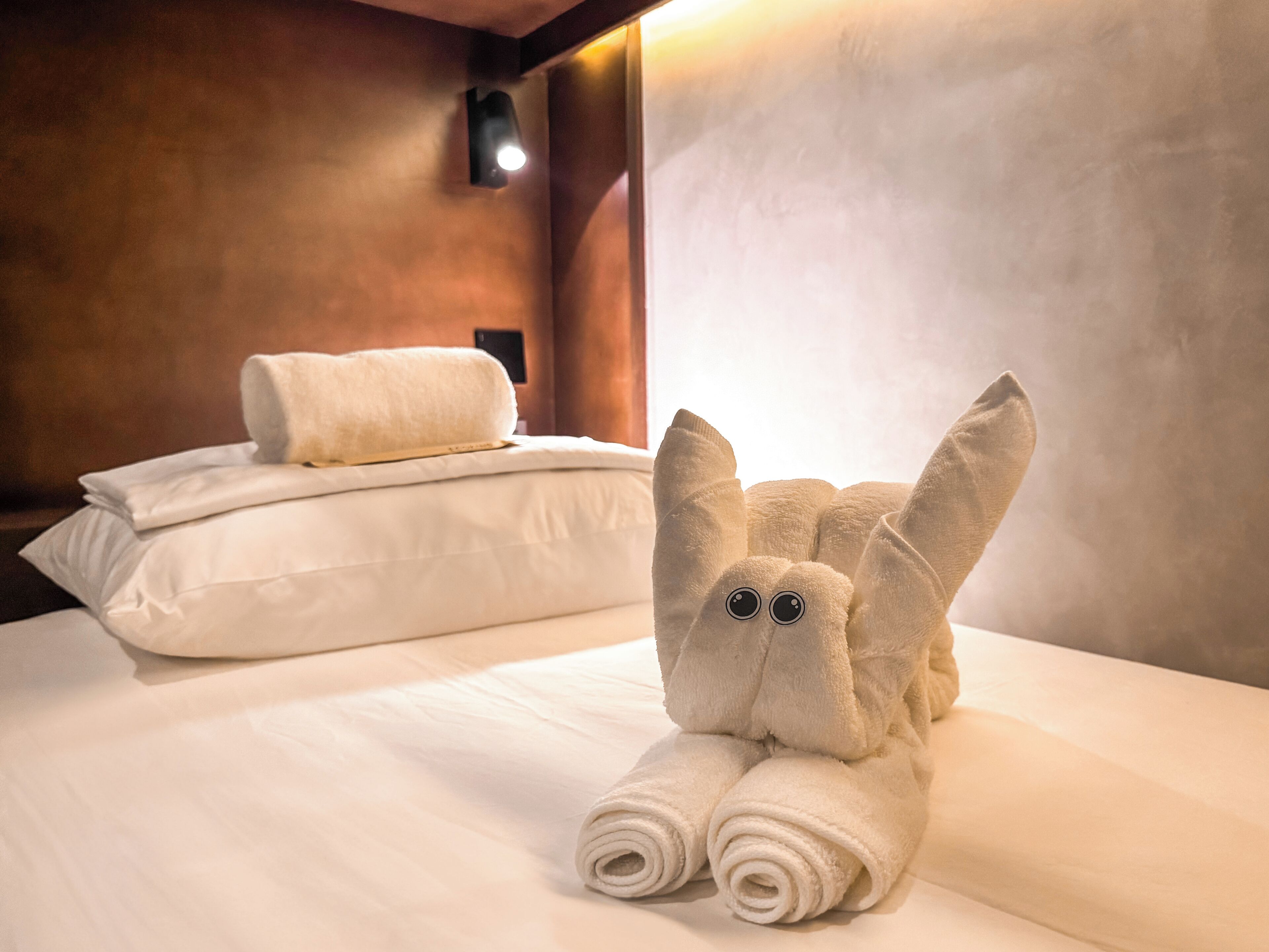 A hotel room with a neatly made bed, a plush toy resembling a rabbit, and a wall-mounted light fixture.