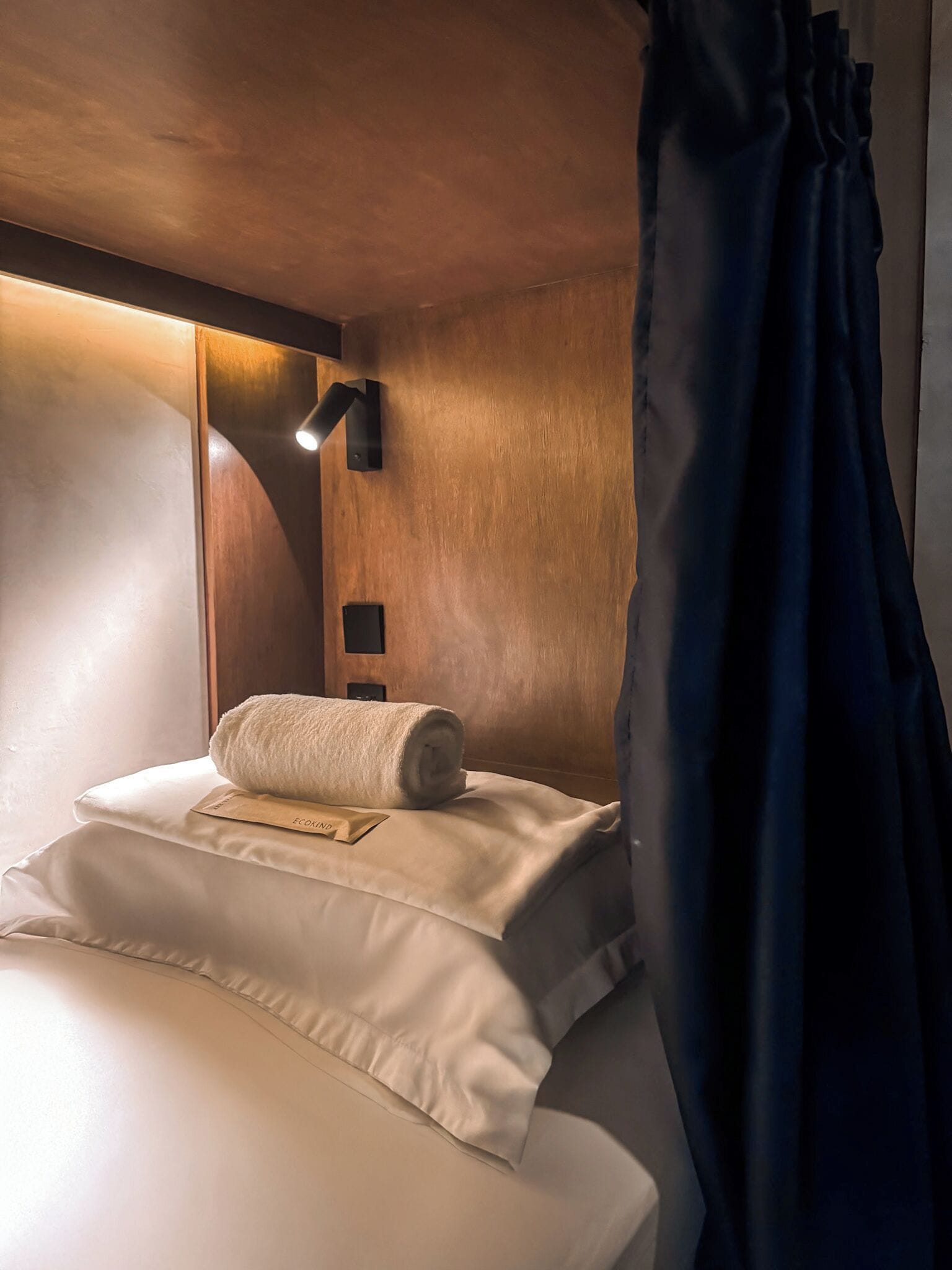 A cozy hotel room with a neatly made bed, a wooden headboard, a wall-mounted reading light, and a dark curtain.