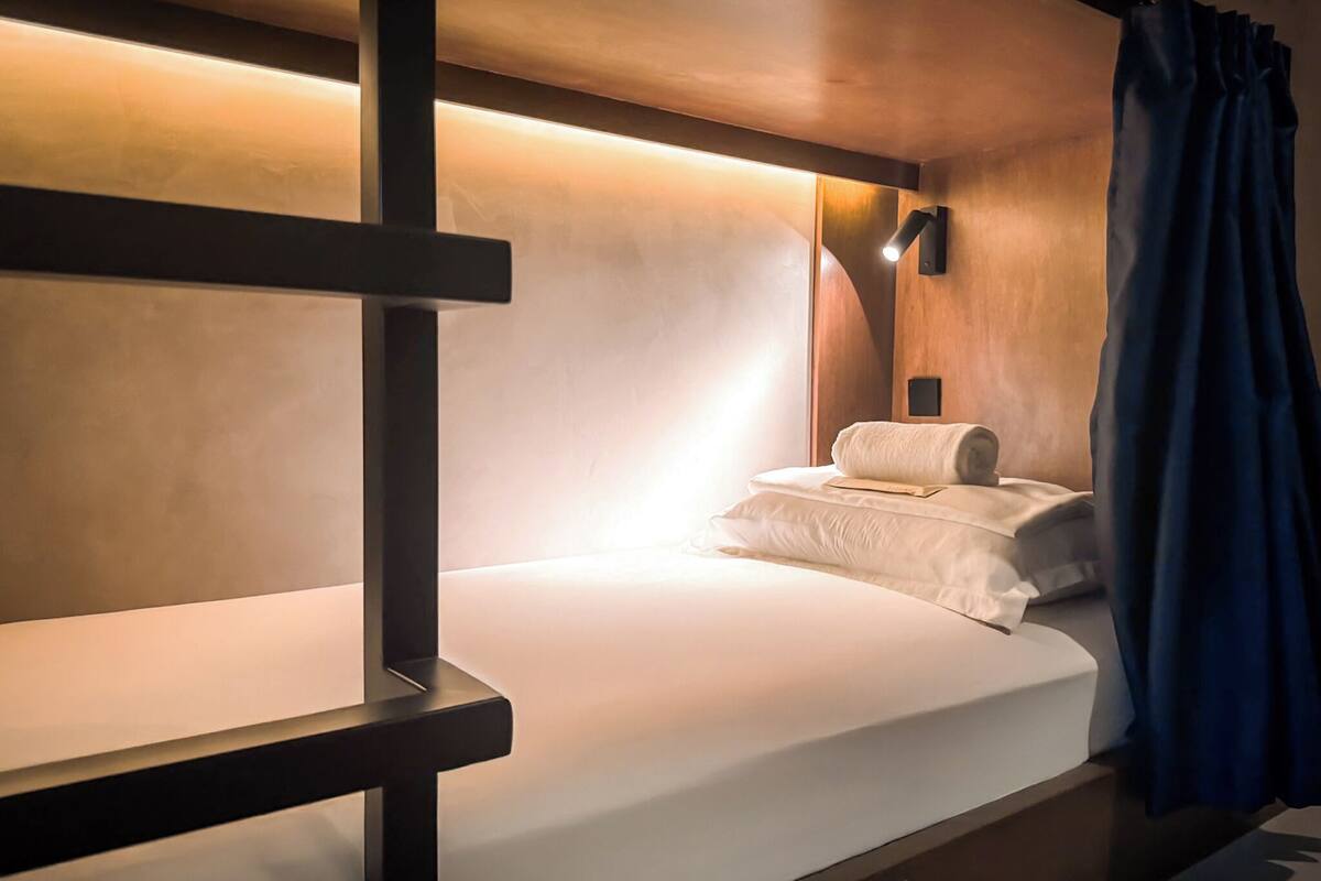A bunk bed with a wooden frame, a white mattress, and a pillow. There is a wall-mounted light fixture above the bed and a curtain on the right side.