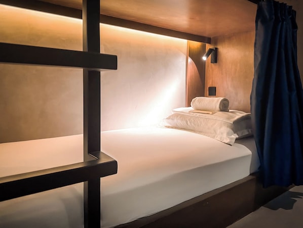 A bunk bed with a wooden frame, a white mattress, and a pillow. There is a wall-mounted light fixture above the bed and a curtain on the right side.