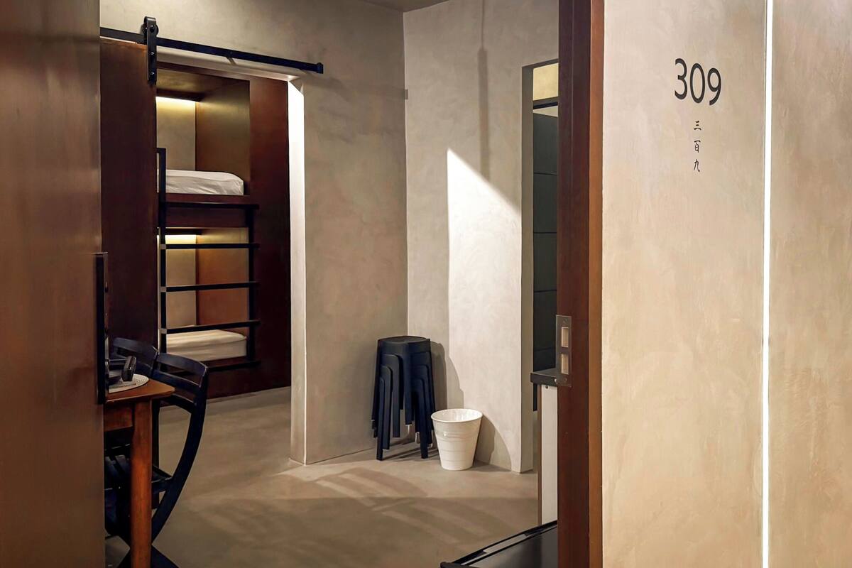 A modern hotel room with a bunk bed, a desk with a chair, a small table, and a trash bin. The room has a minimalist design with a neutral color palette.