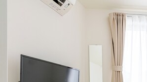 A room with a TV mounted on the wall, a wooden desk, a white chair, a mirror, and a window with curtains.