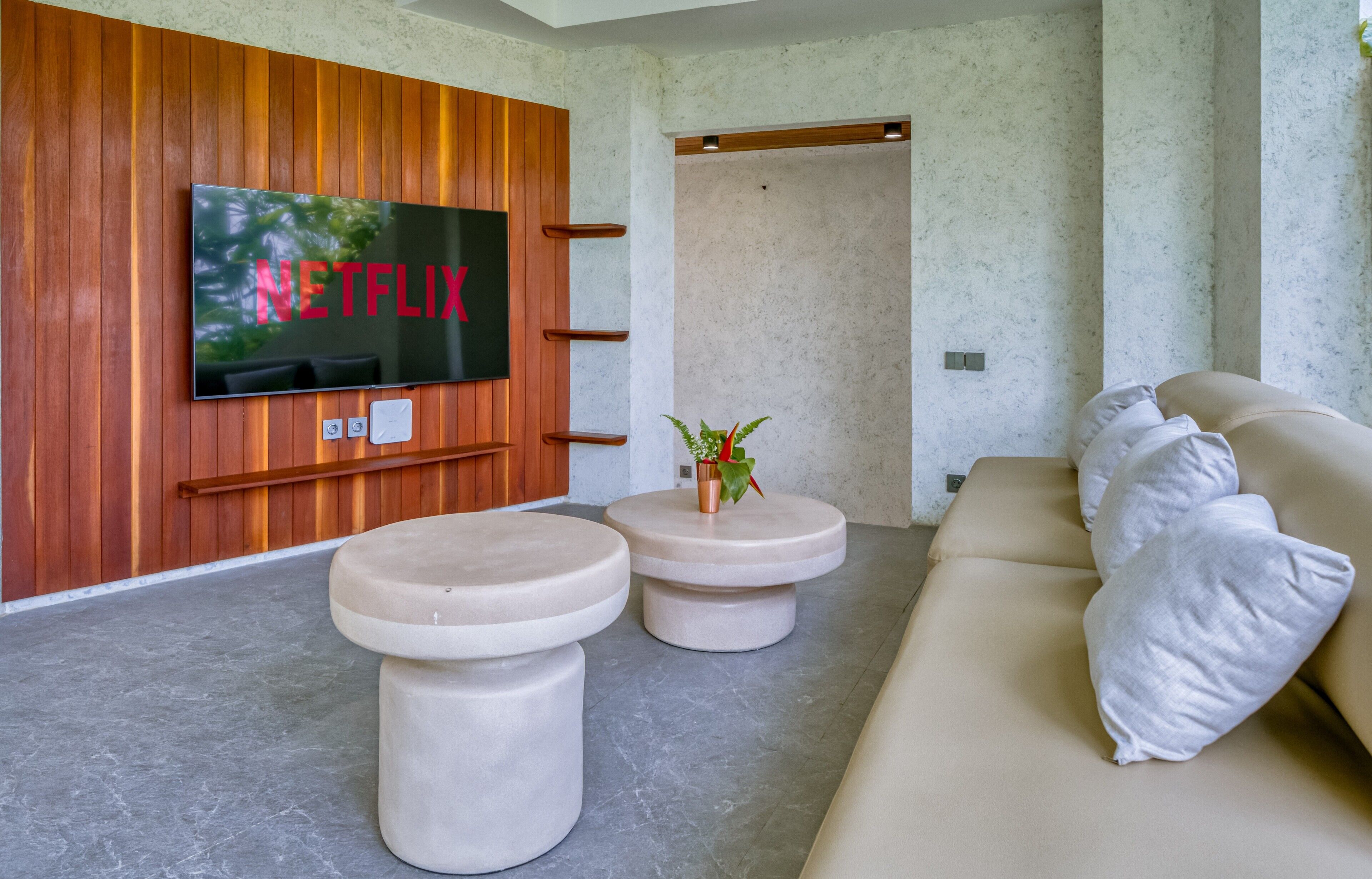 A modern living room with a large flat-screen TV mounted on a wooden wall, two round beige ottomans, a beige sofa with pillows, and a small potted plant.