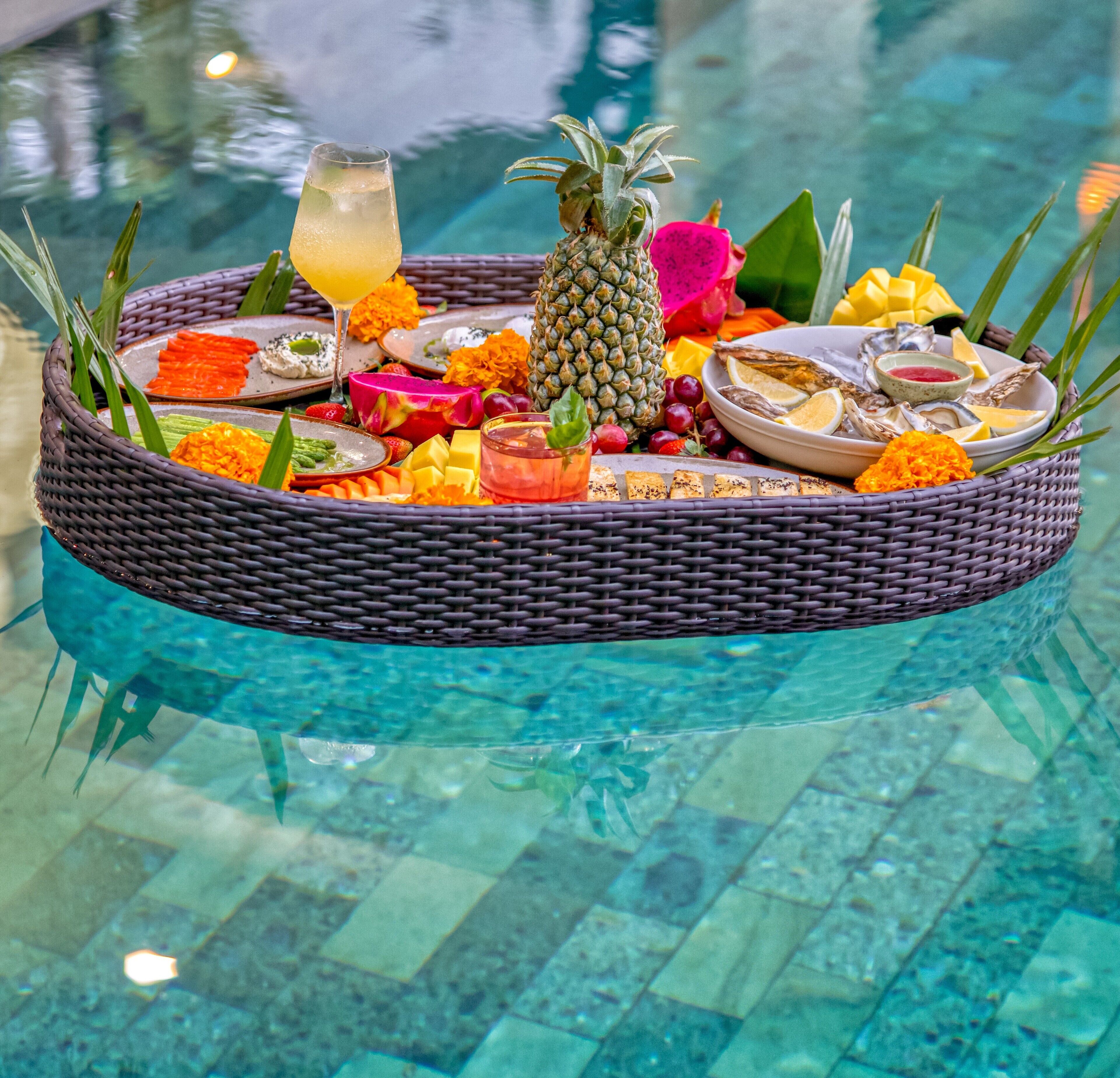 A floating tray with tropical fruits, a drink, and seafood on a poolside.