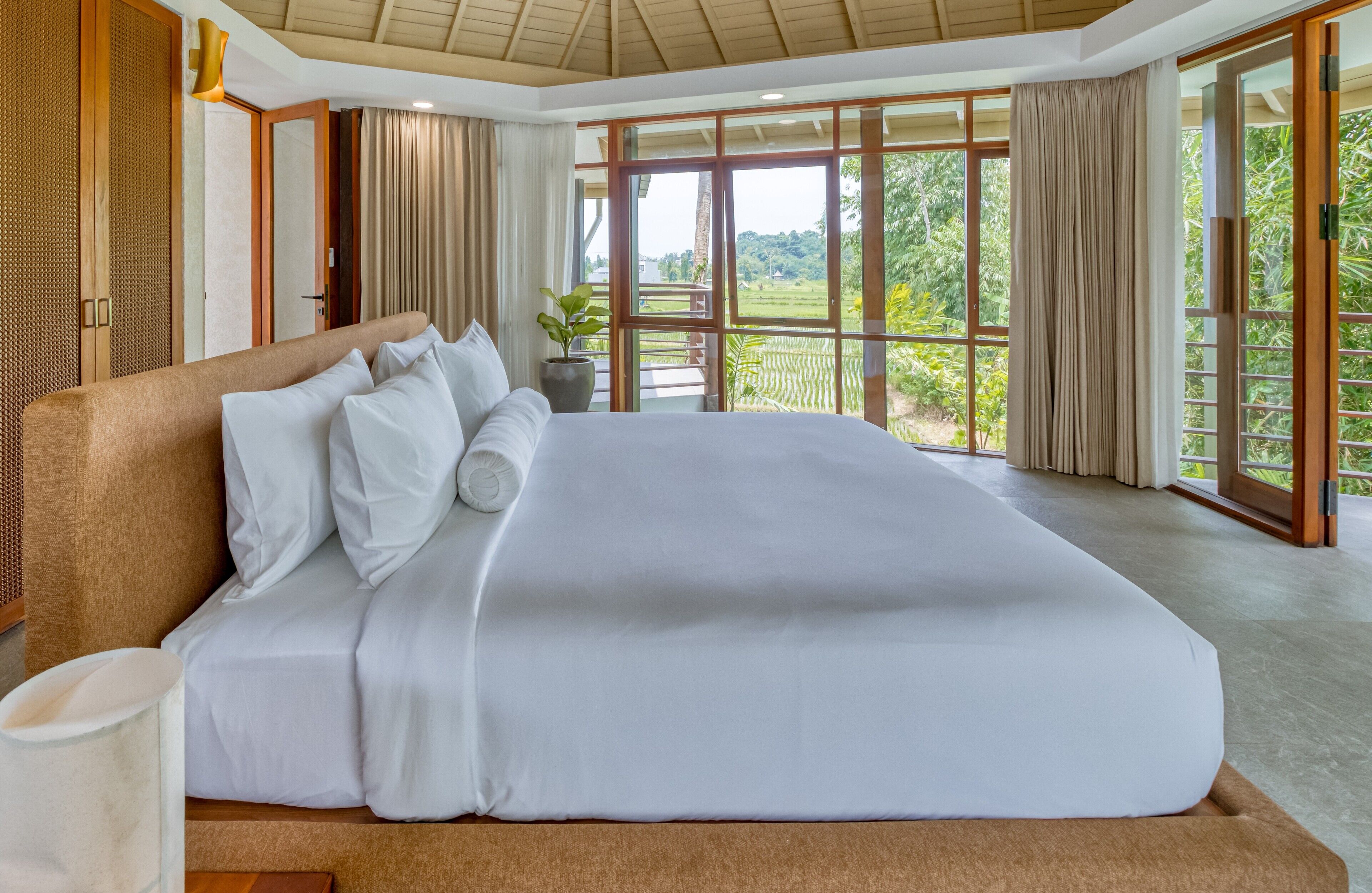 A large bed with white linens in a room with large windows and a view of greenery outside.
