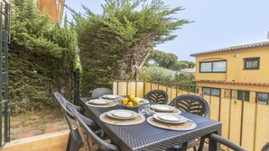 Un espace extérieur aménagé pour les repas, avec une table dressée pour quatre personnes, entourée de chaises en osier, d’une clôture jaune et de verdure.