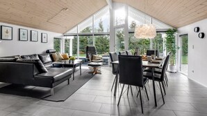 Amplia sala de estar con sofá de cuero negro, mesa de comedor con sillas y grandes ventanales con vistas a la vegetación.