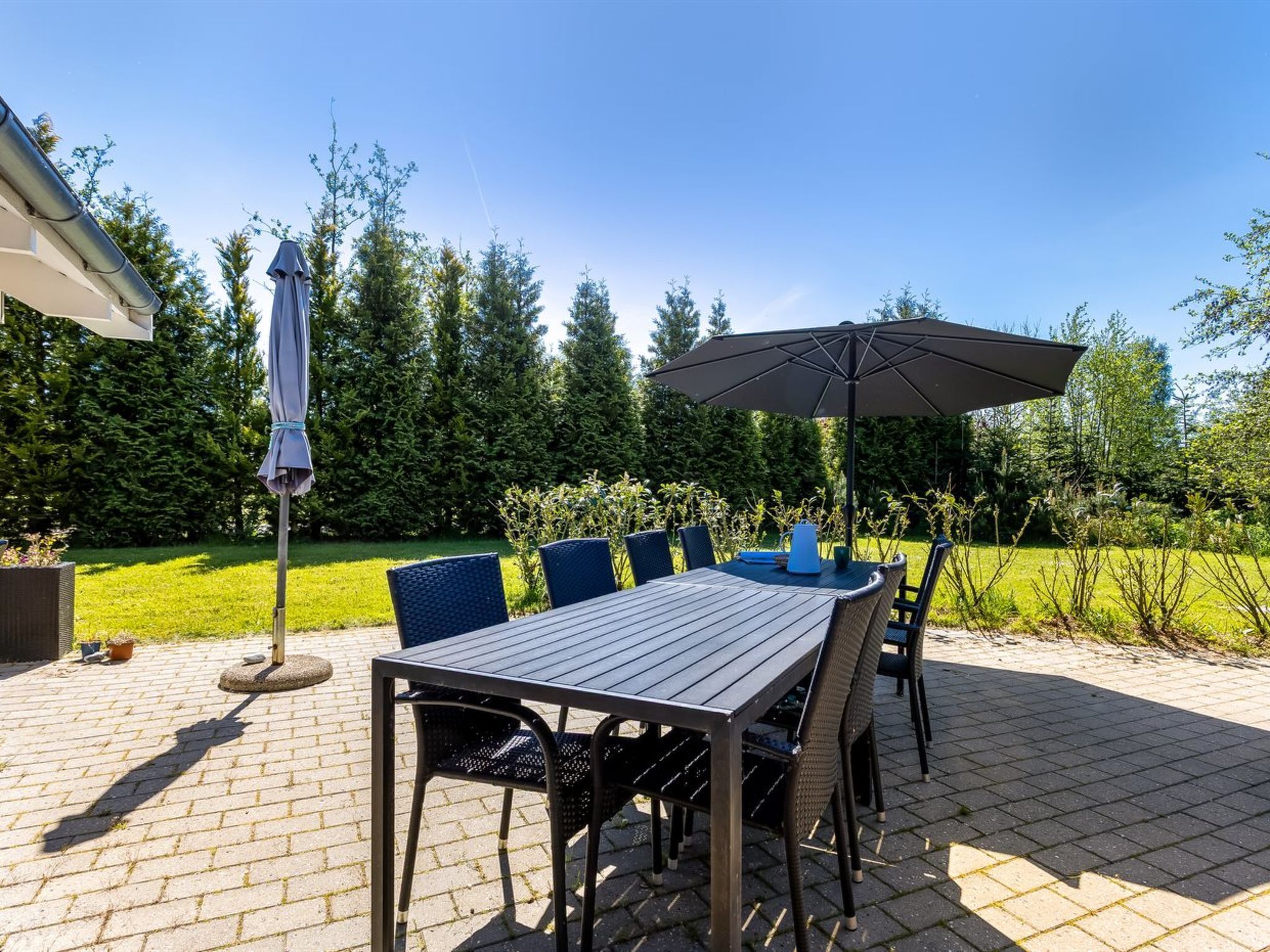 An outdoor patio with a table and chairs, an umbrella, and a garden in the background.
