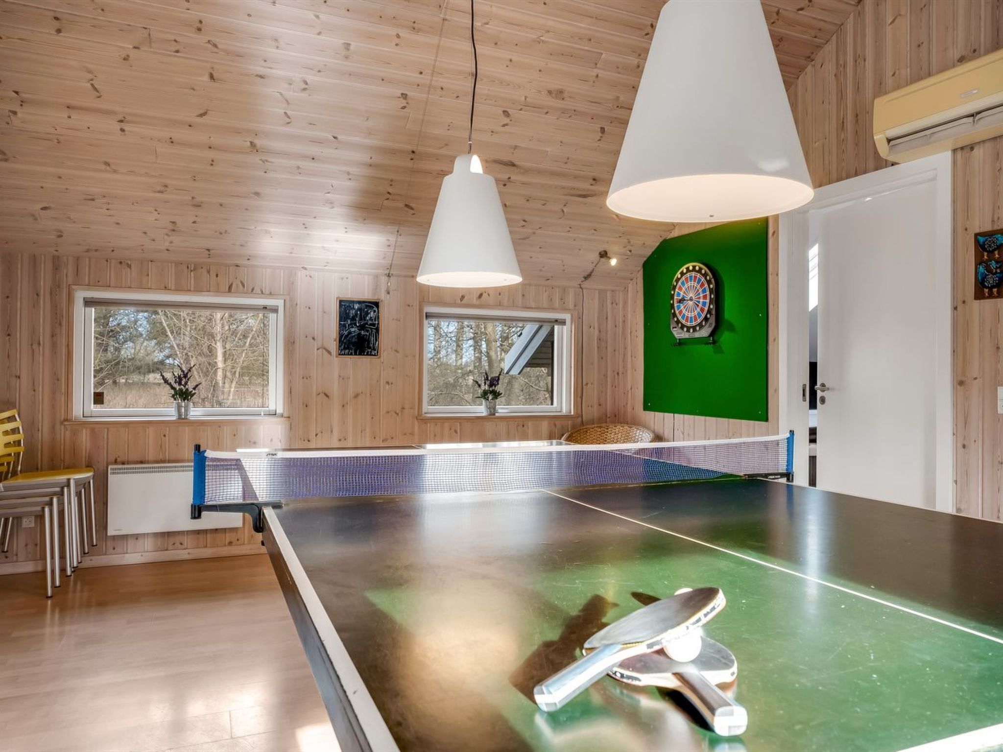 Ein Tischtennistisch in einem Raum mit Holzwänden, großen Fenstern und einer Dartscheibe an der Wand.