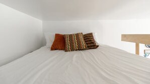 Un lit avec une literie blanche et deux coussins à motifs dans une pièce aux murs blancs et dotée d’une tête de lit en bois.