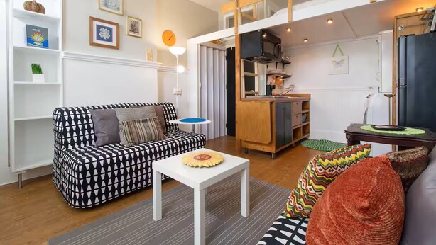 Un salon avec un canapé à motifs, une table basse blanche et un coin cuisine doté de meubles en bois et d’un réfrigérateur.