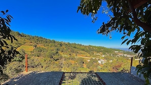 Blick von einer überdachten Außenfläche mit Metallgitterboden auf eine hügelige Landschaft mit verstreuten Gebäuden und Vegetation.