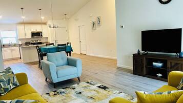 Un salon moderne avec un canapé, une table basse et un téléviseur posé sur un meuble en bois. Au fond, on aperçoit une cuisine avec des meubles blancs et un coin repas comprenant une table et des chaises.